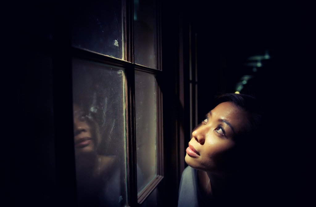 A dark-skinned woman looking out a window toward a night sky. Image by Pexels from Pixabay: https://pixabay.com/photos/alone-dark-female-girl-light-1867056/