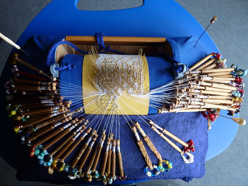 A top-down view photograph of bobbin lace in progress. A card with pins sticking out of it in a lace pattern, dozens of bobbins of thread arrayed around and through the pins to create the patterned lace.