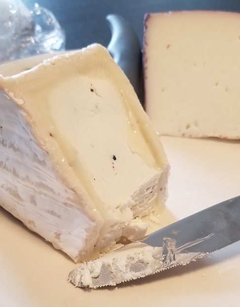 A photograph with two wedges of cheese, the closest of which is a very melty brie that has recently been cut by the dinner knife in the foreground.