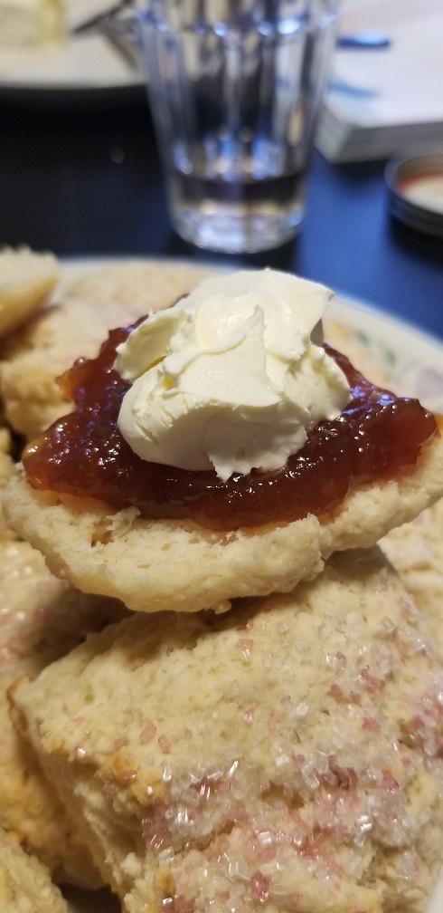 A close-up photograph of a split-open scone with large decorative sugar crystals. One half of the scone is covered with thick red jam and a dollop of clotted cream.