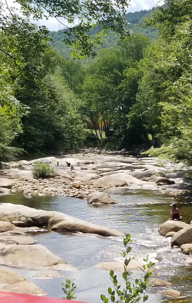 A photograph of a shallow river running over broad, flat rocks, the sun shining brightly over the rippling water. Trees and undergrowth curve up and over the river. A person is sitting on one of the rocks in a swimsuit and hat, their back to the camera. In the distance, a family is playing on the rocks and examining the water pools.