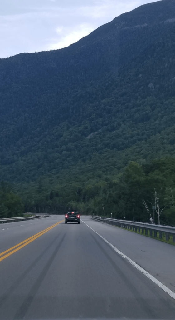 A photograph of the highway through the Notch in the White Mountains of New Hampshire. A single car is braking far ahead; no other cars are on the road that curves out of sight. The highway appears to head straight into a mountain that crowds the rest of the photograph, with only a small triangle of grey-blue sky visible, and no sun.