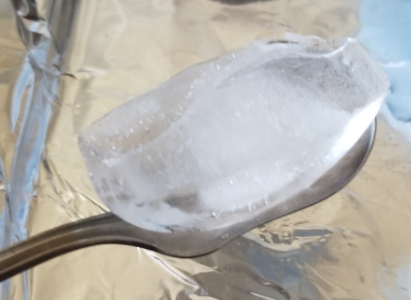 A close-up photo of a silver serving spoon holding a large ice cube.