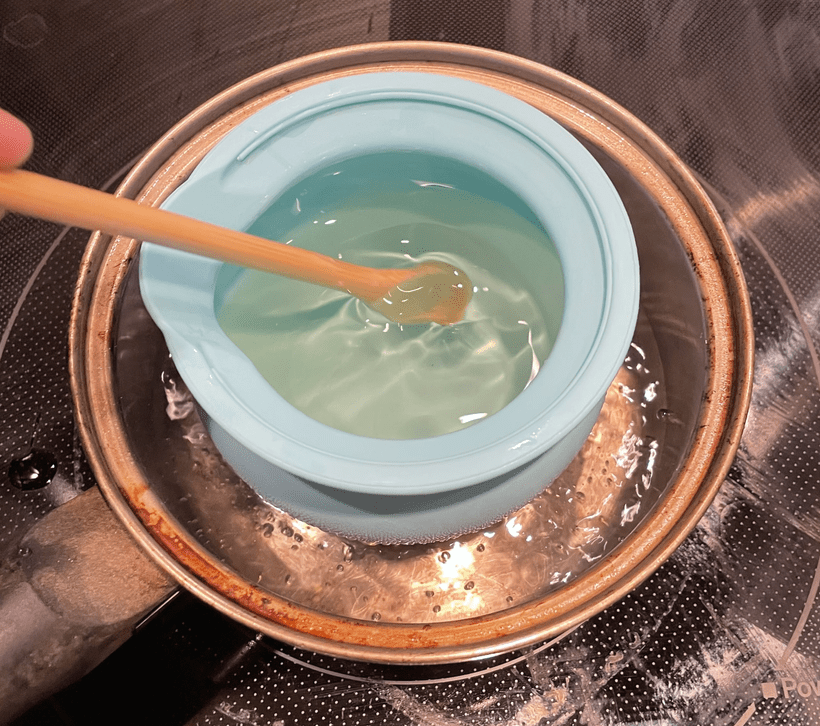 A top-down photograph of a light blue silicone melting pot in a small saucepan of boiling water. A thin wooden spoon is stirring a clear mix of almond oil and beeswax in the melting pot.