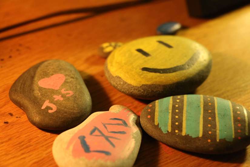 Photograph of several painted stones. One is a yellow happy face; one is a pink heart with "J+S" written on it; one is an Easter-egg-like stripe and dot pattern; and one appears to have Greek lettering on it.