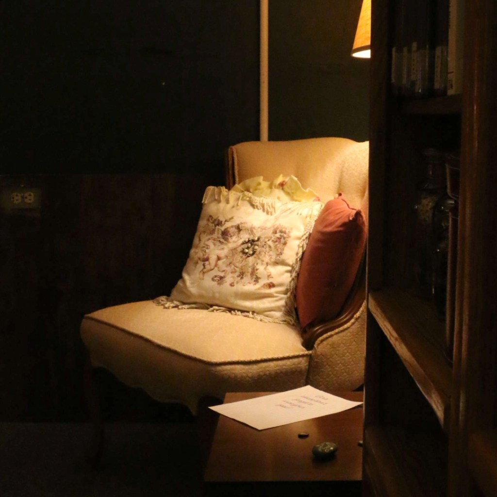 Photograph of an armchair warmly lit in a corner of a dark-green "living room," with a bookcase in the foreground.