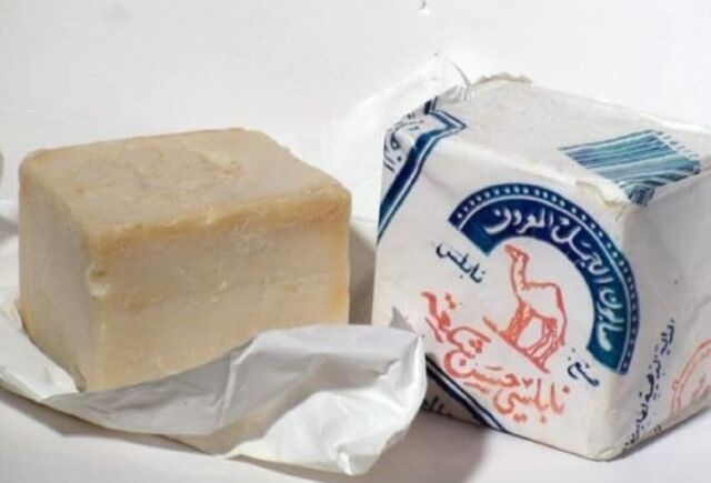 A close up of stock photo of a wrapped and an unwrapped pair of Nabulsi soaps. The unwrapped soap is irregularly cut and slightly off-white, with the look of something hand-cut (because it is); the wrapped soap shows the blue and white company information, with a red camel in the center.
