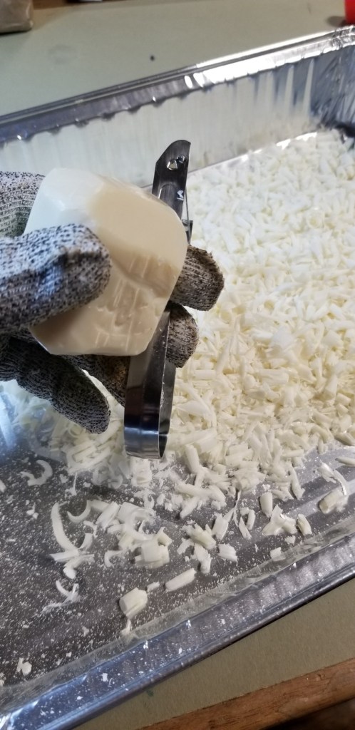Fairly close-up photo of a large disposable cake tin with a great deal of curled up slivers of peeled soap in it. In the foreground, a hand covered in a metal-mesh safety glove holds a partially denuded bar of soap and a potato peeler.