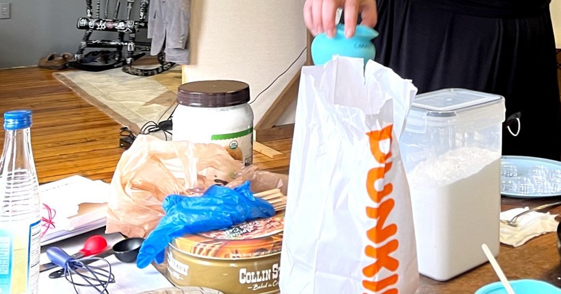 A close-up photograph of a Dunkin Donuts bag on a table of lip-balm production tools and ingredients.