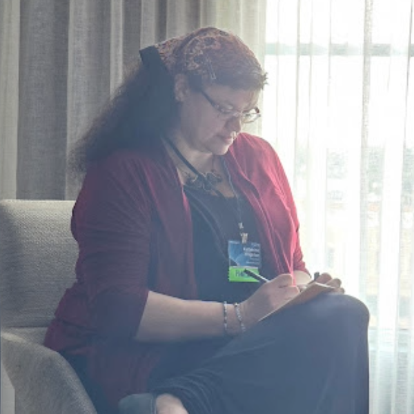 Photograph of Katherine Crighton sitting in a chair in a hotel room, focused on writing in a small notebook. They are wearing a panelist badge for Readercon 2024.