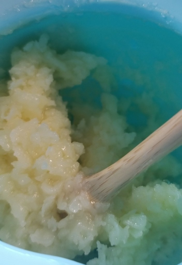 Photograph of the inside of a blue silicone pot containing a yellow-ish white mixture of oil, beeswax, and rosewater, which is starting to form clumps that look like cottage cheese around a small wooden spatula