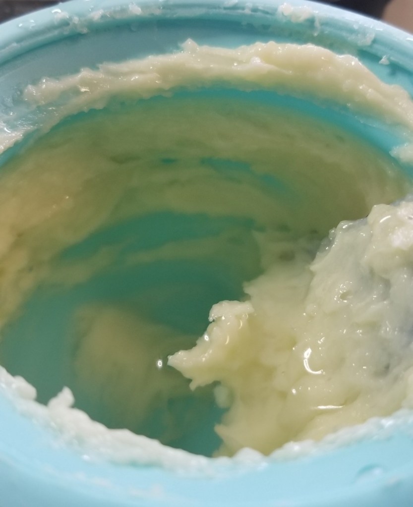 Photograph of the inside of a blue silicone pot containing a yellow-ish white mixture of oil, beeswax, and rosewater, which is starting to smooth out into something the consistency of whipped frosting around a small wooden spatula.