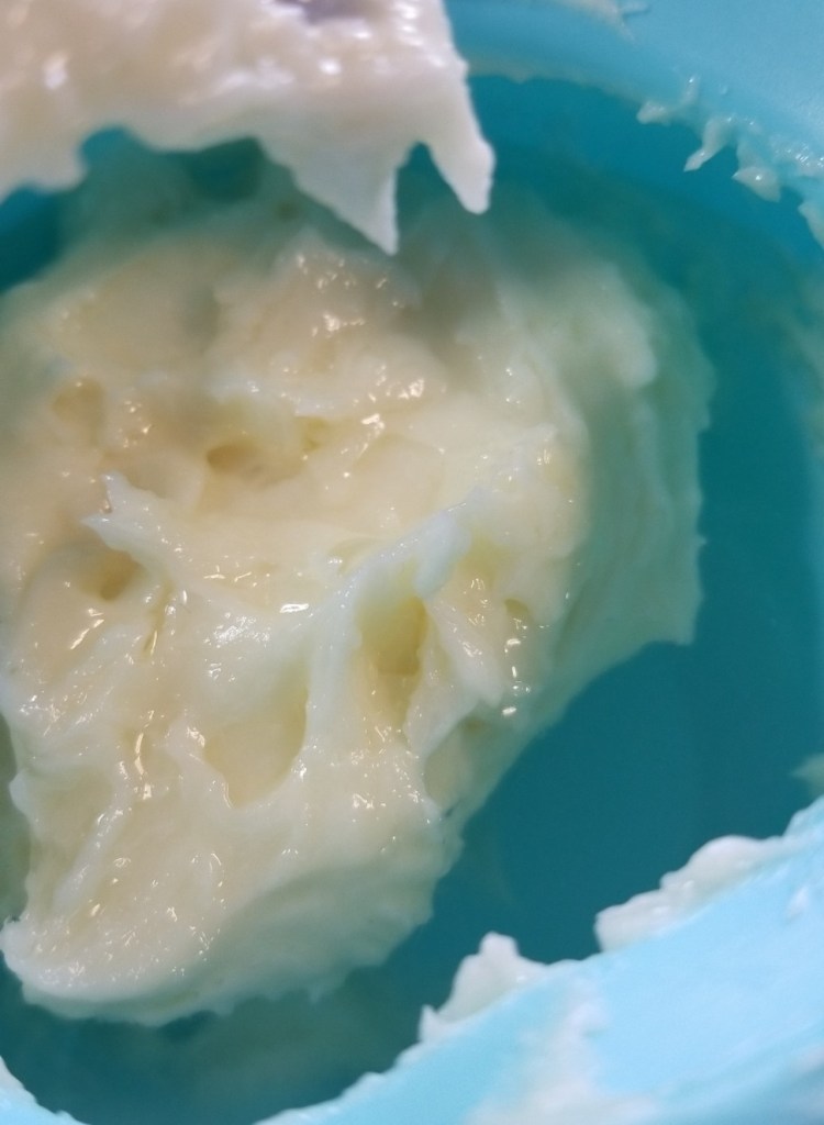 Photograph of the inside of a blue silicone pot containing a white-ish mixture of oil, beeswax, and rosewater, which appears to look like a thick lotion and has been formed into a rough sphere, floating in the rosewater.