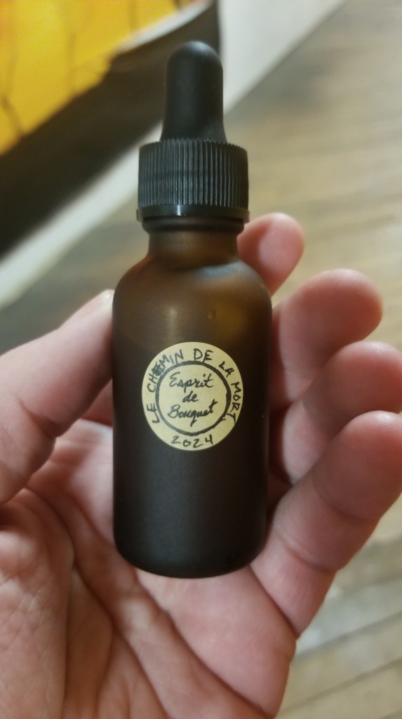 Close-up photograph of a dark brown, matte glass bottle with a black dropper and a small circular kraft-paper label with "LE CHEMIN DE LA MORT 2024: Esprit de Bouquet" handwritten on it.
