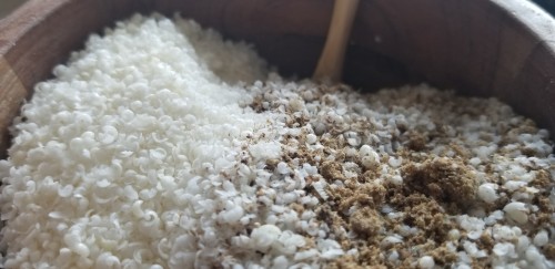 Photograph of tiny curls of shaved, dried white soap partially mixed with a brown powder in a wooden mortar, the long, thin handle of a wooden spoon sticking out of it.