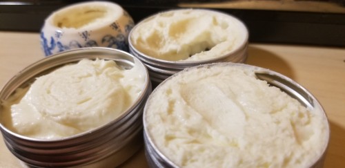 Close-up photograph of three metal tins and a small ceramic pot holding a thick, white, creamy pomatum for the skin, the recipe of which has been later described as a cold cream. The pomatum has the appearance of whipped butter.