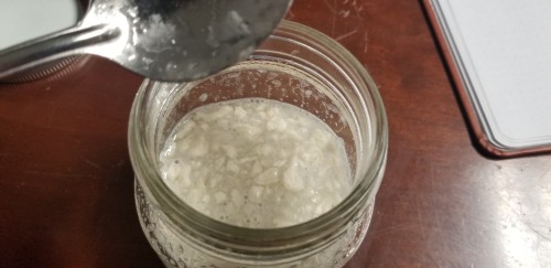 Overhead photograph of the inside of a small glass jar, a dirtied spoon blurry in the foreground. The inside of the jar has a liquid mess of oatmeal-colored water and distressing clumps.