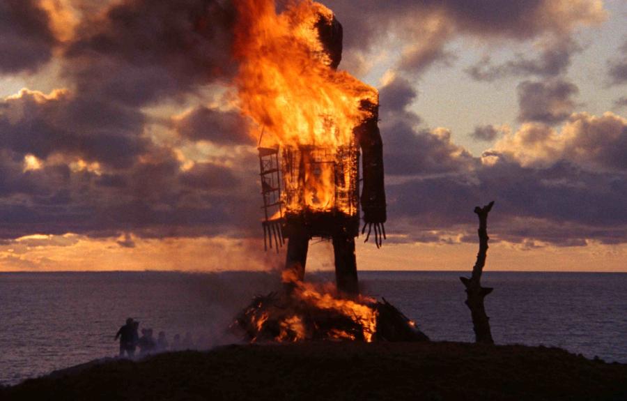 Publicity still from The Wicker Man (1973). A beachhead in silhouette, with some shadowy figures and a stubby tree, against an orange, cloud-filled sunset, frames the center figure: an enormous wood and straw construct of a man set aflame, the fire raging across its chest and at its feet, surely killing any sassenach coppers stuffed inside it.