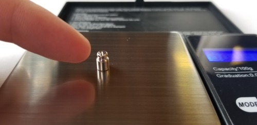 Close-up photograph of a finger pointing at a 1-gram calibration weight on a digital scale. It is incredibly tiny.