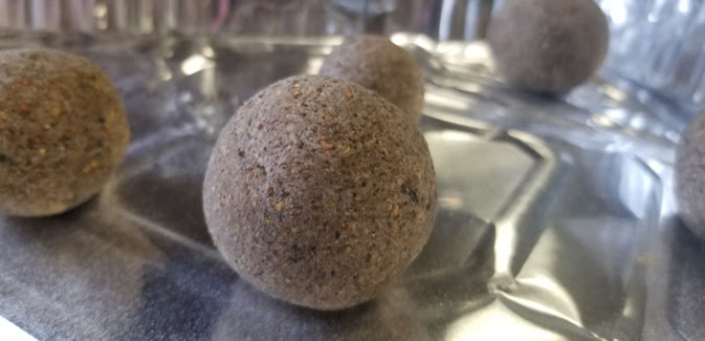 Close up photograph of a speckled, smooth sphere of soap, green-grey with very small dots of dark brown and black evenly interspersed, appearing very like granite or similar igneous rock. In the background are three other soaps of a similar size, and one smaller one.