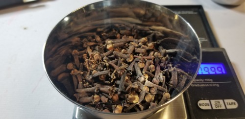 Photograph of a metal small bowl full of whole cloves on a digital scale.
