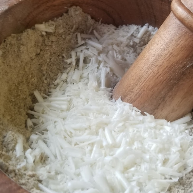 Close up of the bowl of a large wooden mortar filled with green-grey powder and, on top of that, a great deal of shaved and dried white soap (some in pencil-shaving sized curled, some grated more finely with a microplane grater). A wooden pestle is nestled in the pile, about to start mixing.