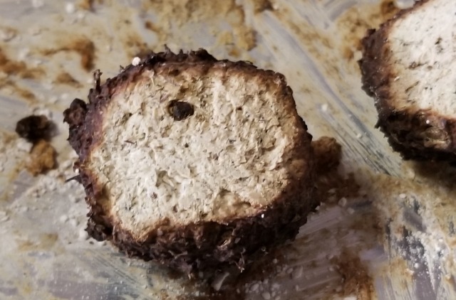 A closeup photograph of a sheer cross-section of a larger one of the extremely brown, knobbly, fecal-looking dry spheres. The cross-section shows that the brown "rind" penetrates a couple of millimeters, and from there the interior looks more like oatmeal or, perhaps, nougat with some ground vanilla bean. The background is a metal tray that appears to have had the mixture that made up these balls poorly scraped off at some point in the past, so that in actuality it appears as if someone took a shit in the pan and then tried (badly) to wipe it up. NONE of this helps the overall impression of this soap.