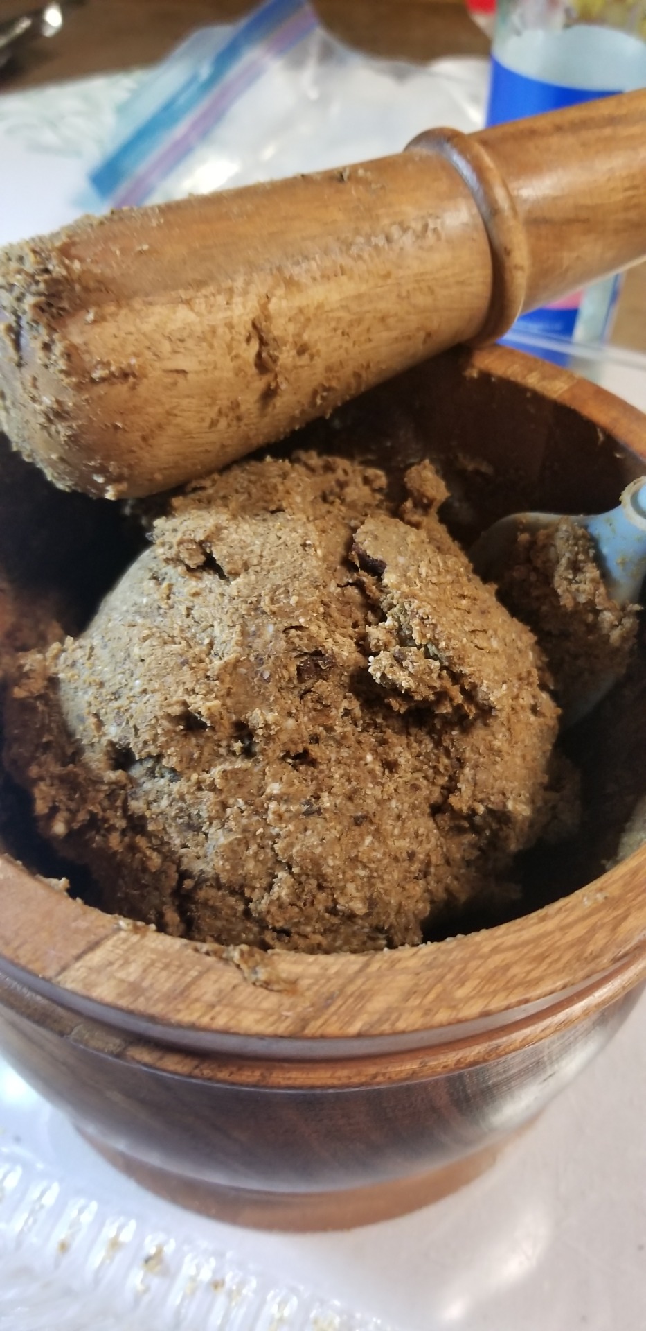 A close-up photograph of a rough, light brown ball of what appears to be dough, but is actually a scented soap mix, in a wooden mortar. A wooden pestle is resting on the mortar, with clumps of the mix sticking to it. Barely visible is a spatula standing upright in the mortar. In the background is a bottle of rosewater, partially obscured.