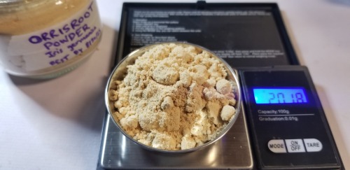Photograph of a bowl of yellow-ish, clumping powder (ground orrisroot) on a small digital scale. Behind it is a glass jar hand-labeled "ORRISROOT POWDER" with the scientific name and the best-by date beneath it.
