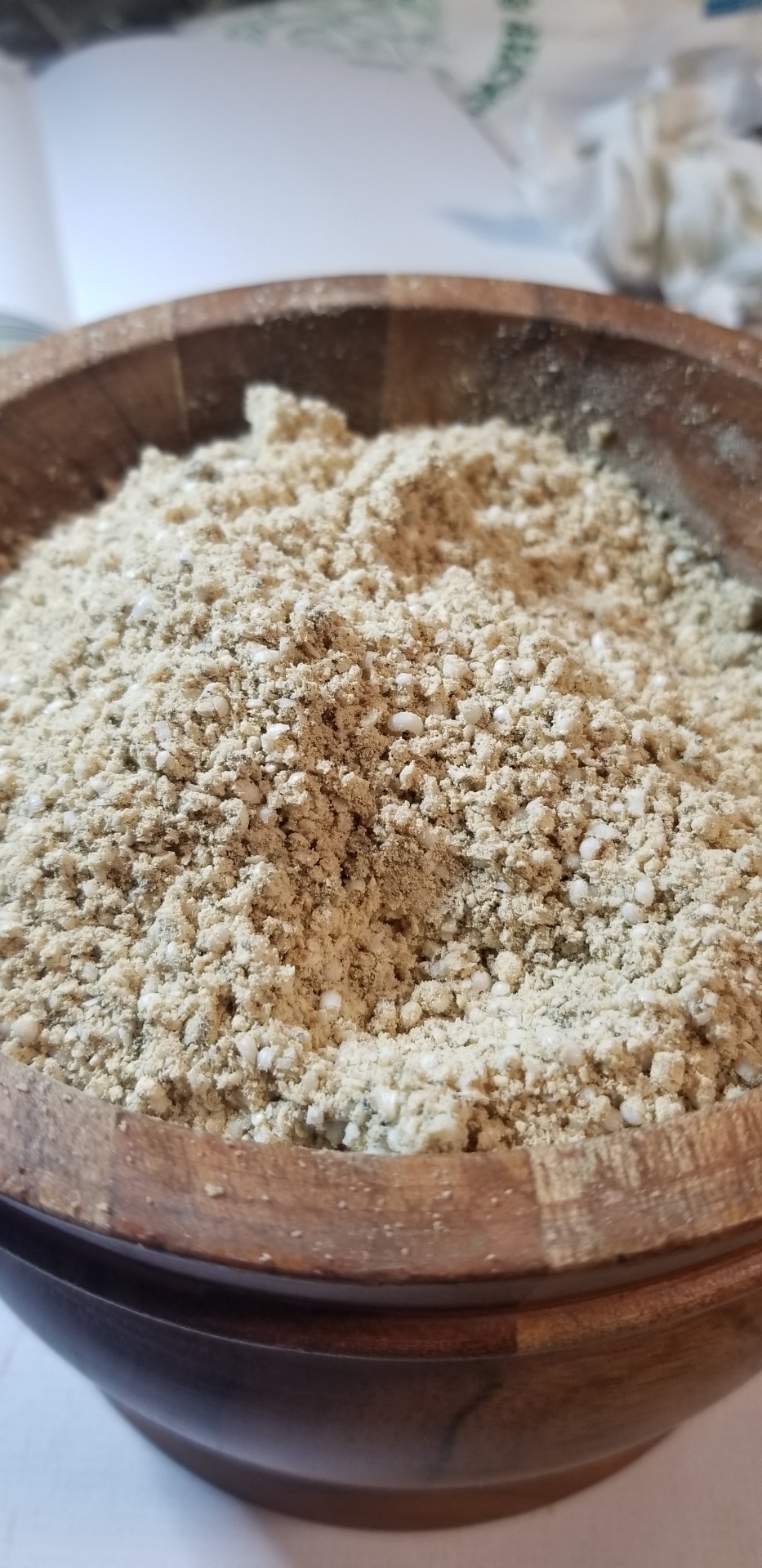 A close-up photograph of a wooden mortar very very full of a mix of ground or powdered ingredients and dried, grated soap. The color is a sort of pleasant light wheatish.