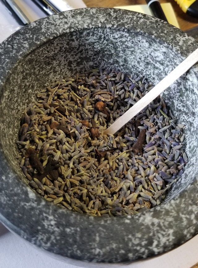 Close up photograph of a granite mortar with 1 oz of whole cloves and a large handful of dried French lavender flowers in it. A wooden stirrer is sticking out of it; the cloves and lavender have been lightly stirred so both are visible for the camera.